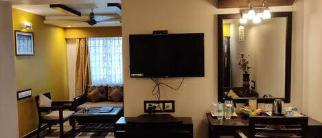 A hotel room with a flat-screen TV mounted on the wall, a dining table with chairs, a sofa, and a small table with a mirror and decorative items.