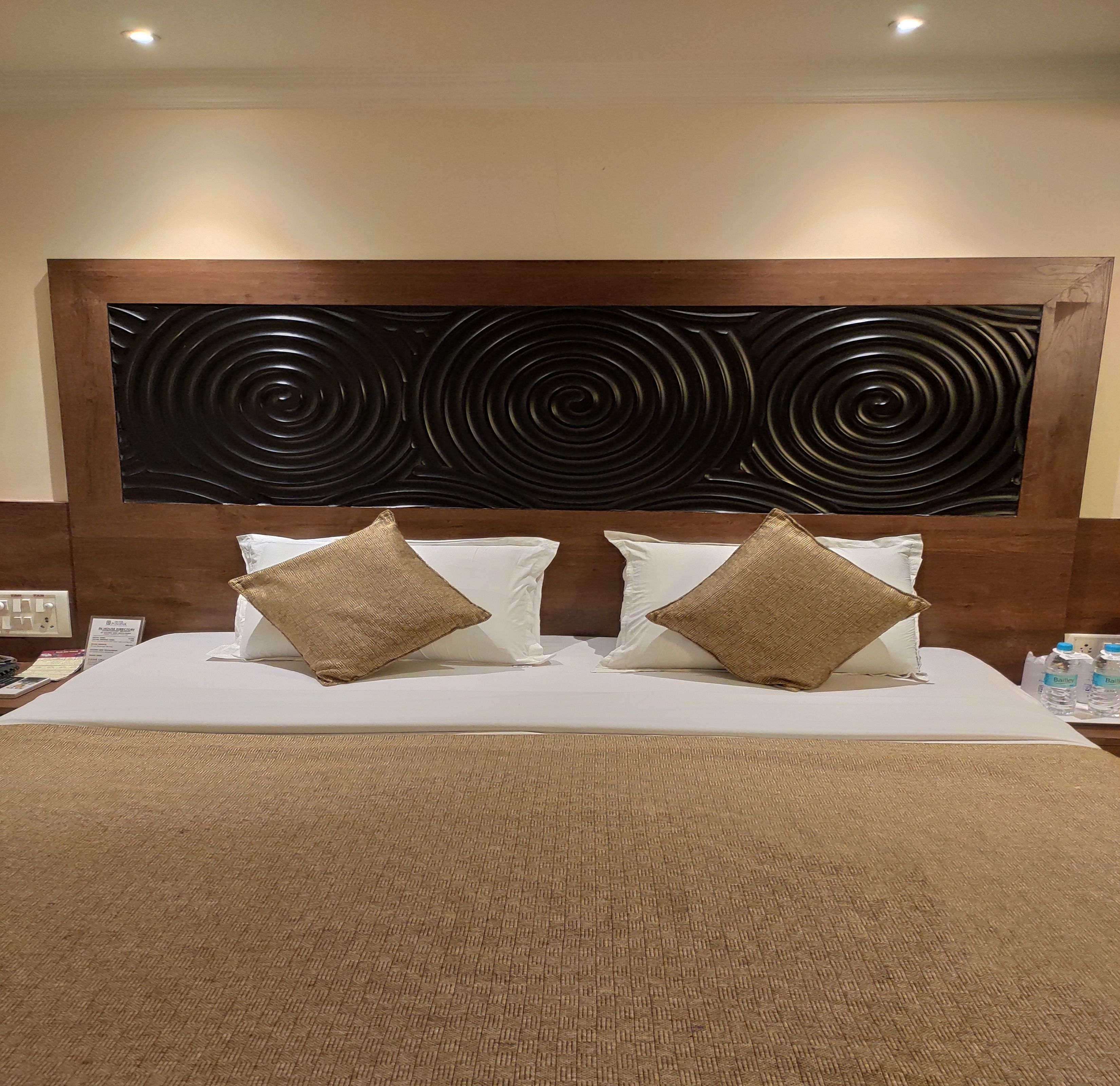 Una habitación de hotel con una cama grande, dos almohadas, un cabecero con diseño circular negro y una alfombra marrón con textura.