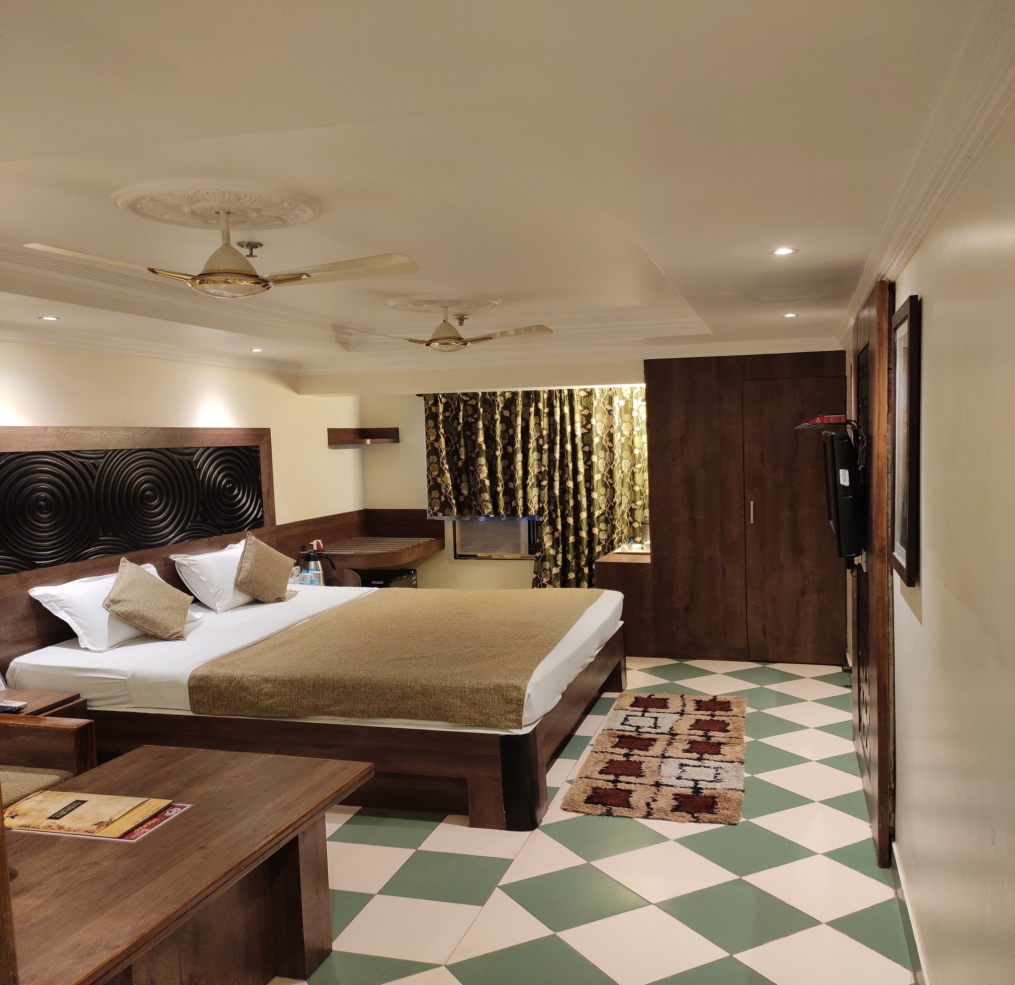 A hotel room with a bed, bedside tables, a desk, a chair, a TV, a ceiling fan, a patterned curtain, and a checkered floor.