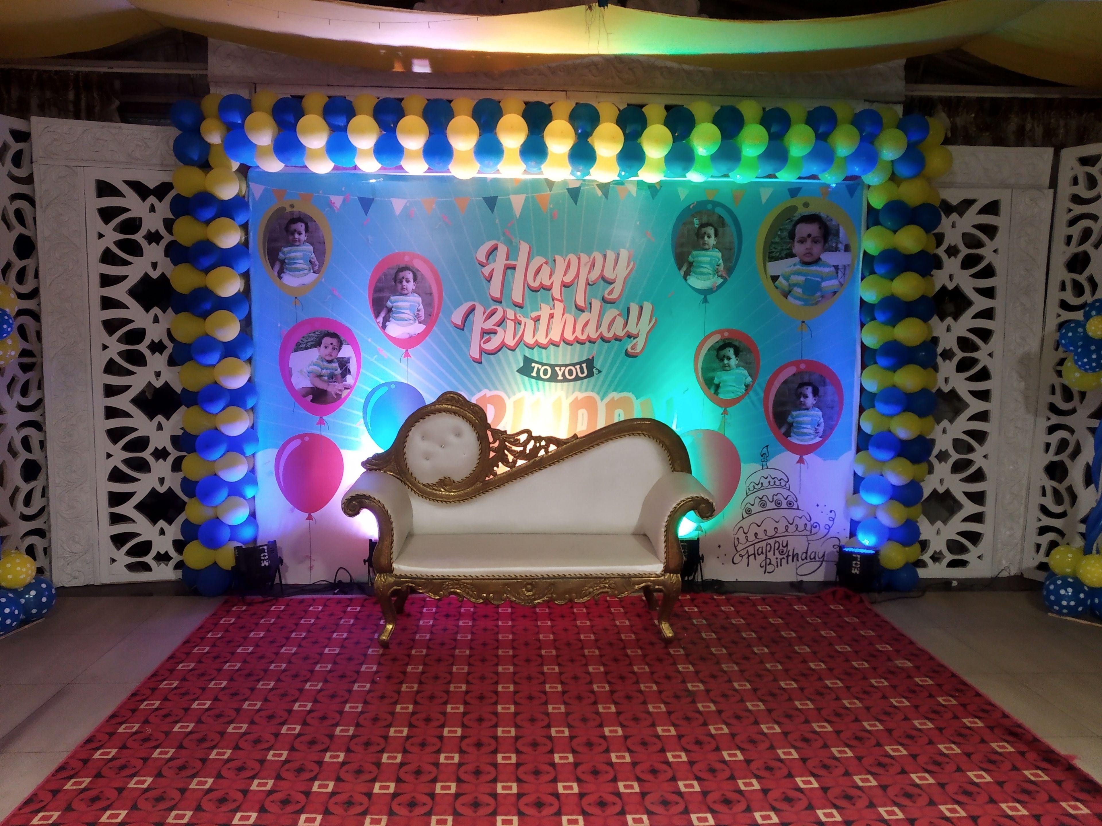 Un escenario con alfombra roja estampada, un sofá blanco y un fondo decorado con el texto 'Happy Birthday' y globos.