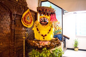 A statue of a deity with a yellow robe and a decorative umbrella, placed on a rock base with plants around it.