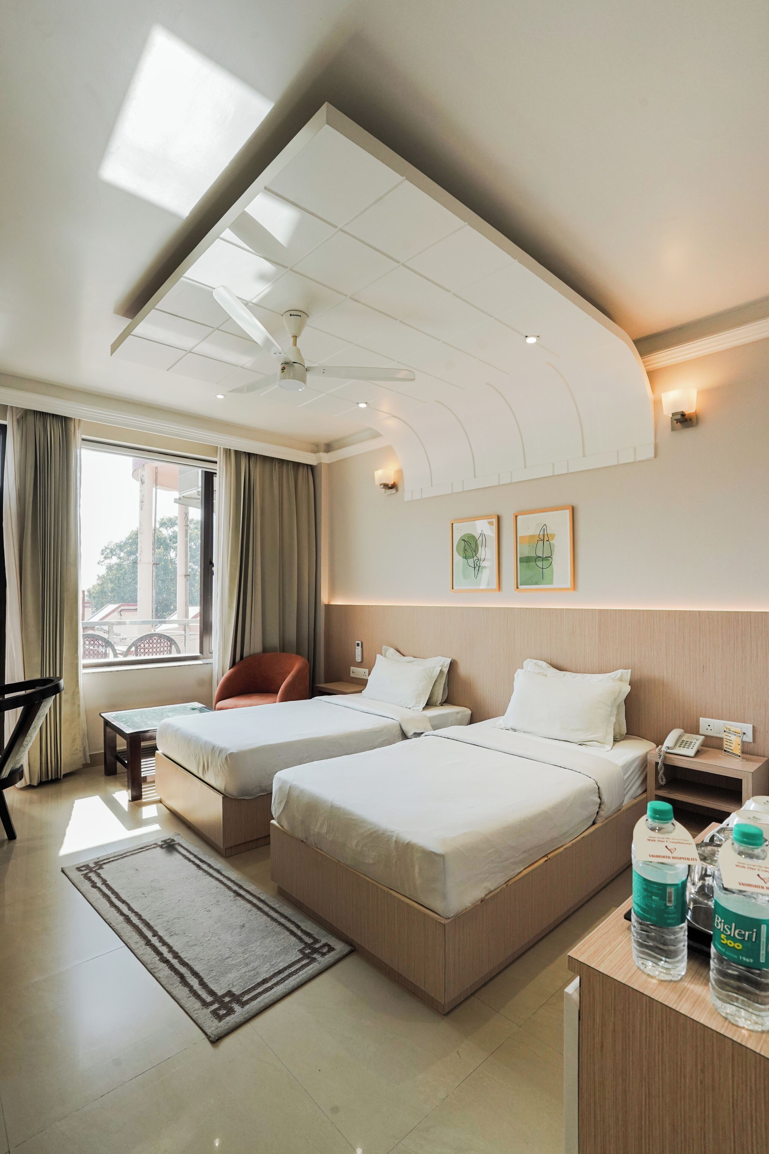 A hotel room with two beds, a desk, a chair, a window with curtains, a ceiling fan, and artwork on the wall.