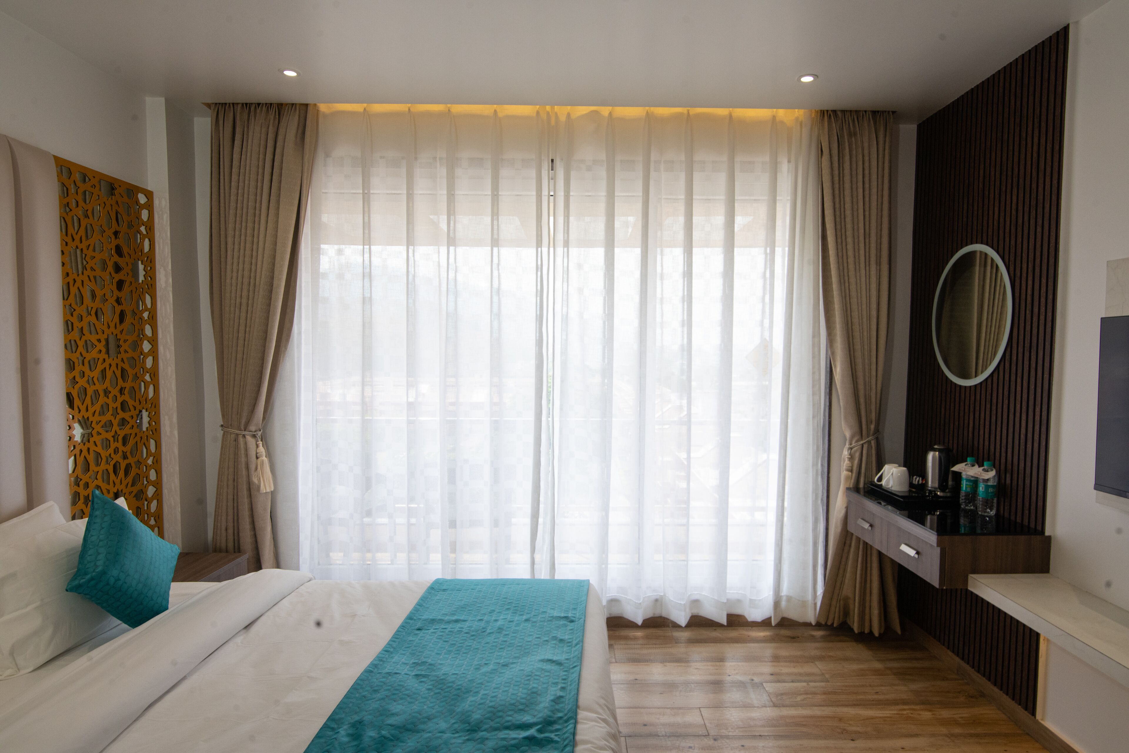 Quarto de hotel moderno com cama grande, piso de madeira, escrivaninha embutida com espelho e uma janela ampla com cortinas leves.