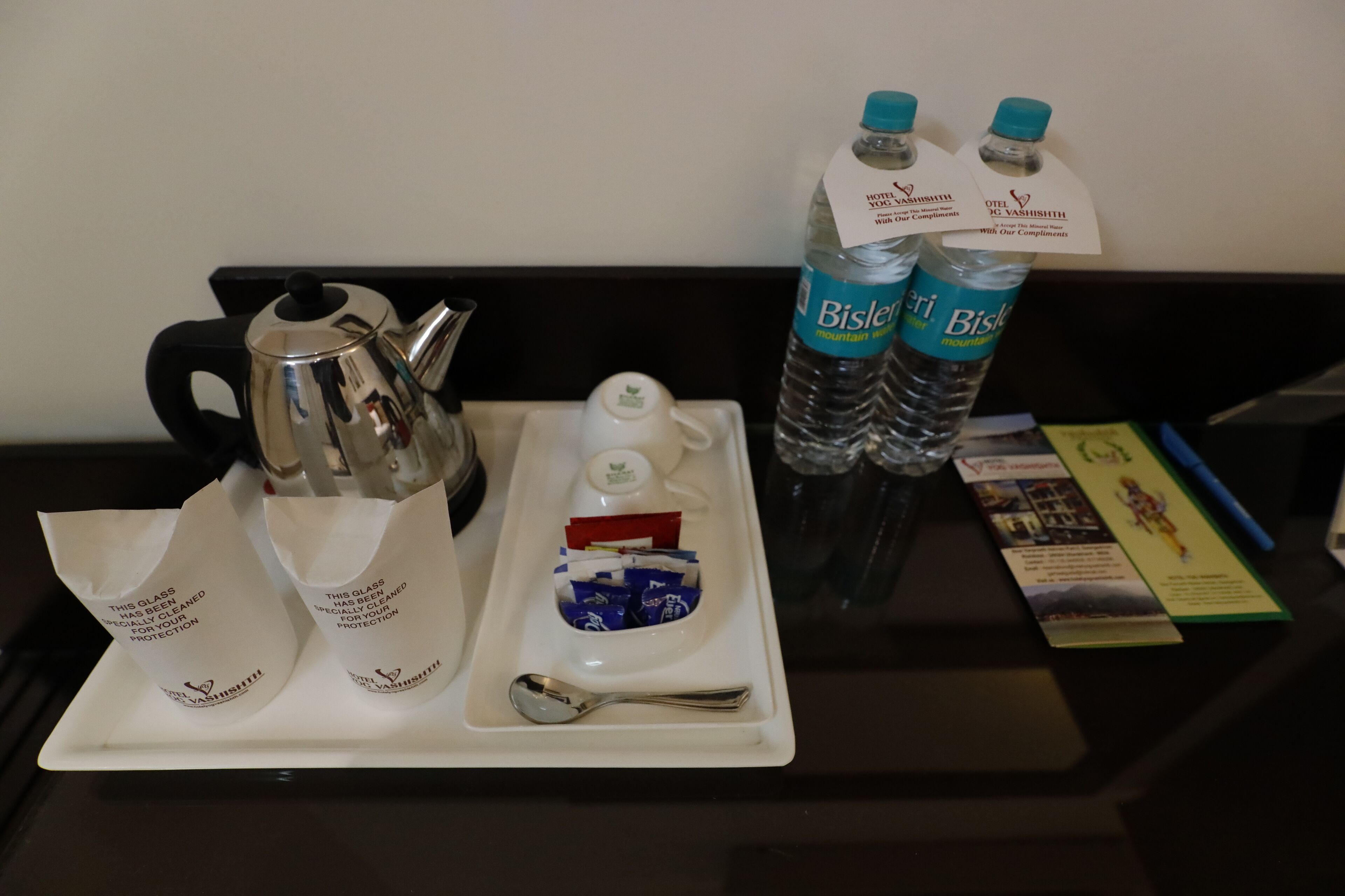Een dienblad met een koffiepot, twee Bisleri waterflessen, een lepel en een klein schaaltje met verpakkingen, alles op een tafel.