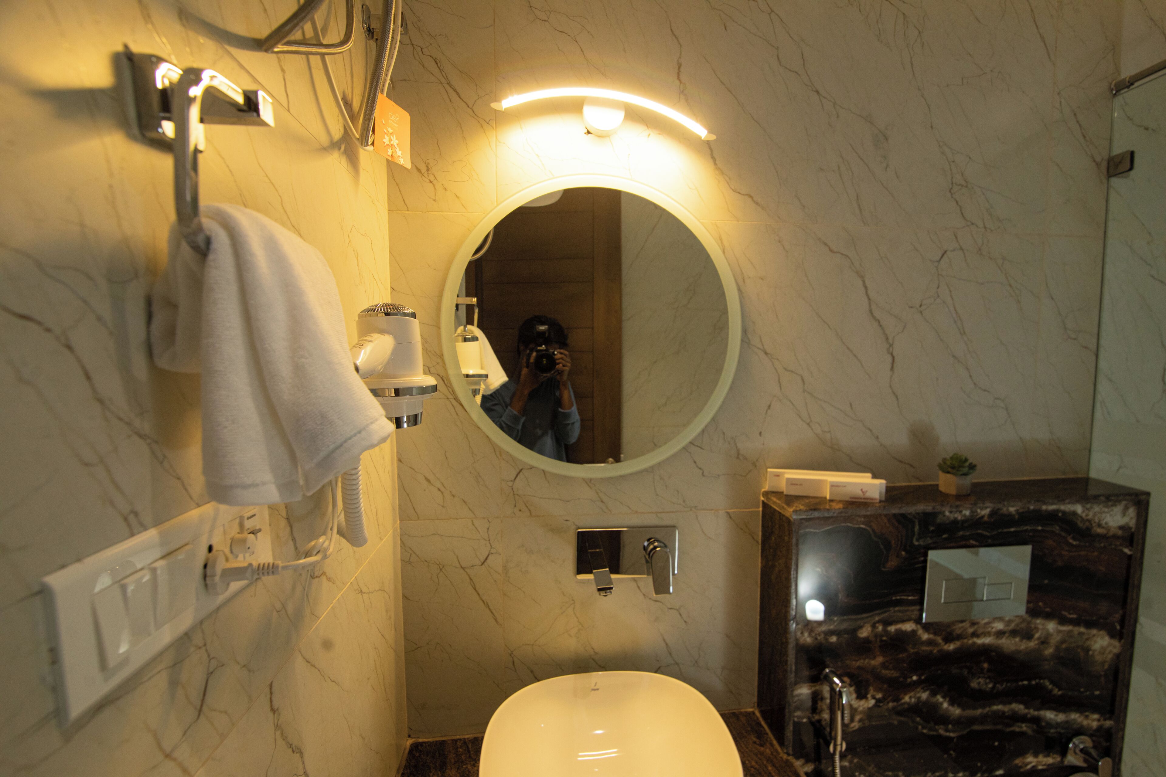 A bathroom with a marble wall, a round mirror, a white sink, and a towel rack.