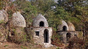 A row of stone buildings with arched doorways and windows, surrounded by trees and vegetation. - Hotel Yog Vashishth (Yamkeshwar)