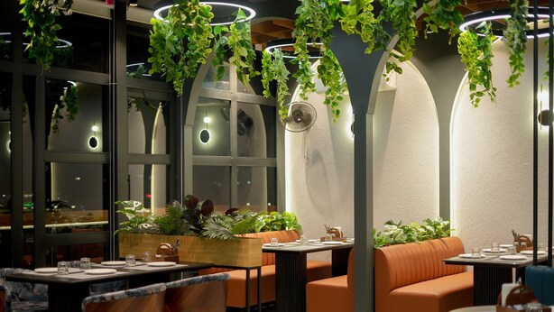 A restaurant with booths and tables, indoor plants, and a patterned floor. - Hotel Yog Vashishth (Yamkeshwar)