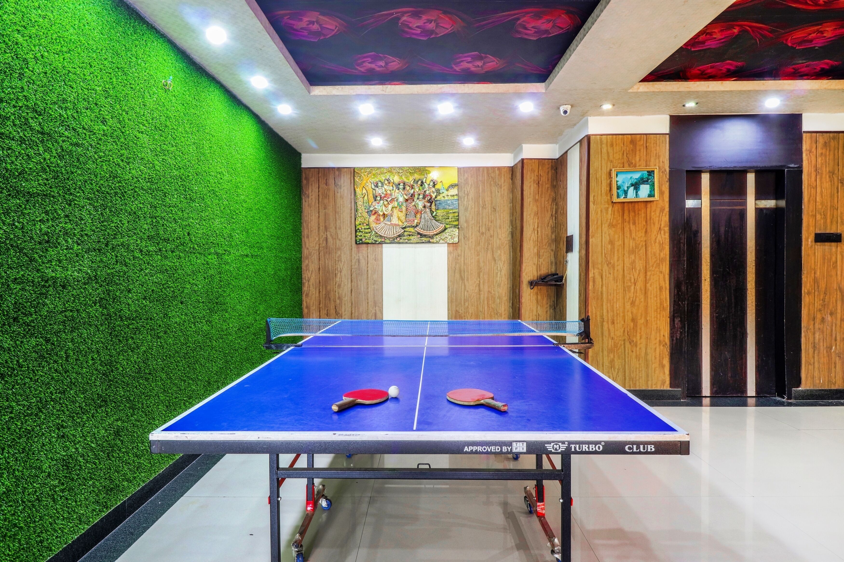Una mesa de ping pong en una habitación con paredes de césped artificial verde, paneles de madera y un techo con estampados florales rojos.