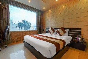 A hotel room with a large bed, a window with curtains, a nightstand with a coffee pot, and a view of palm trees and buildings. - Hotel Grand Shiva (Haridwar)
