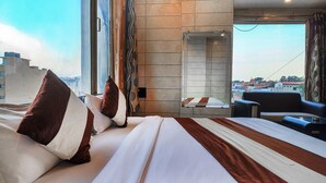 A hotel room with a large bed, a seating area with a sofa and armchairs, a view of the cityscape, and a large window with curtains. - Hotel Grand Shiva (Haridwar)