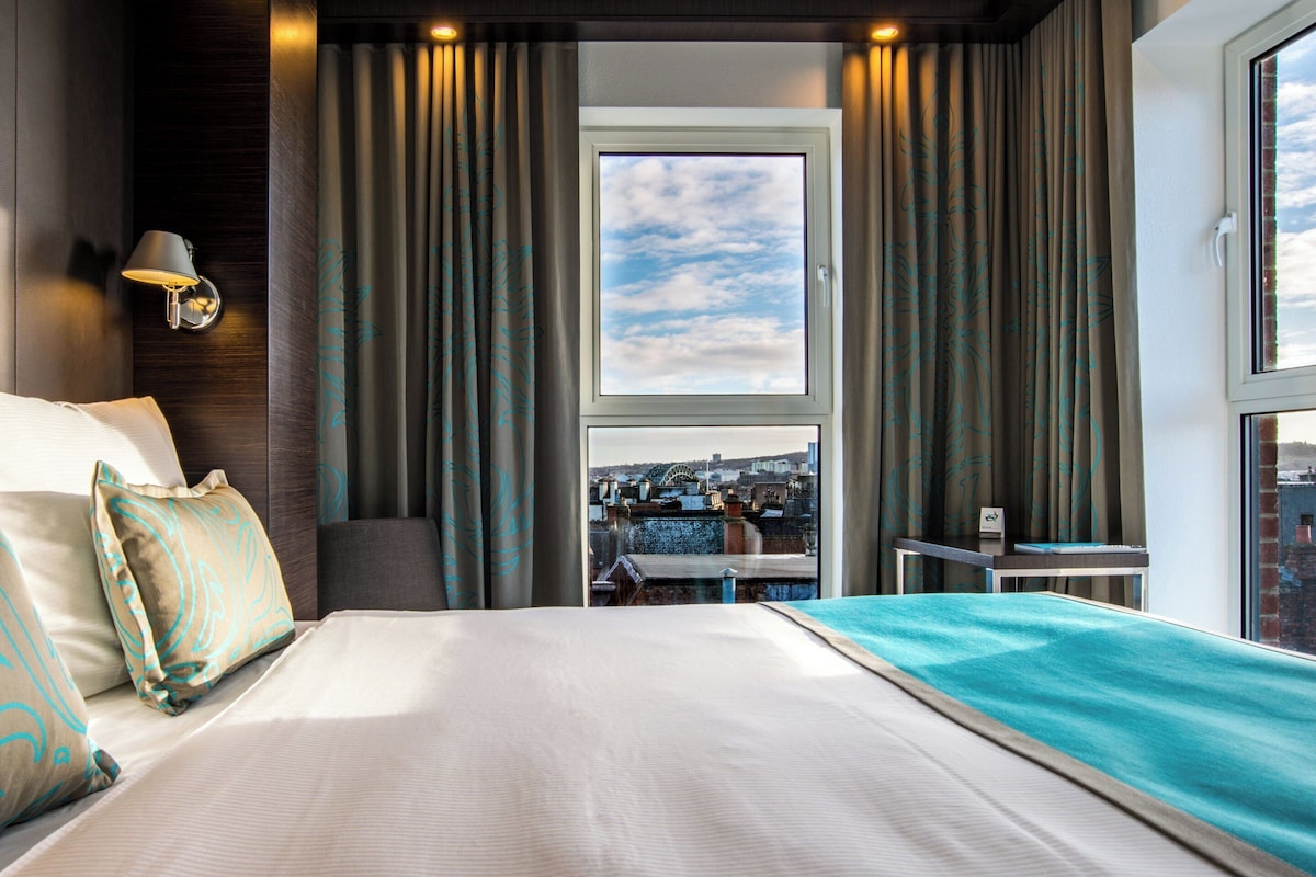 A hotel room with a large bed, a view of the city through a window, and a desk with a chair.
