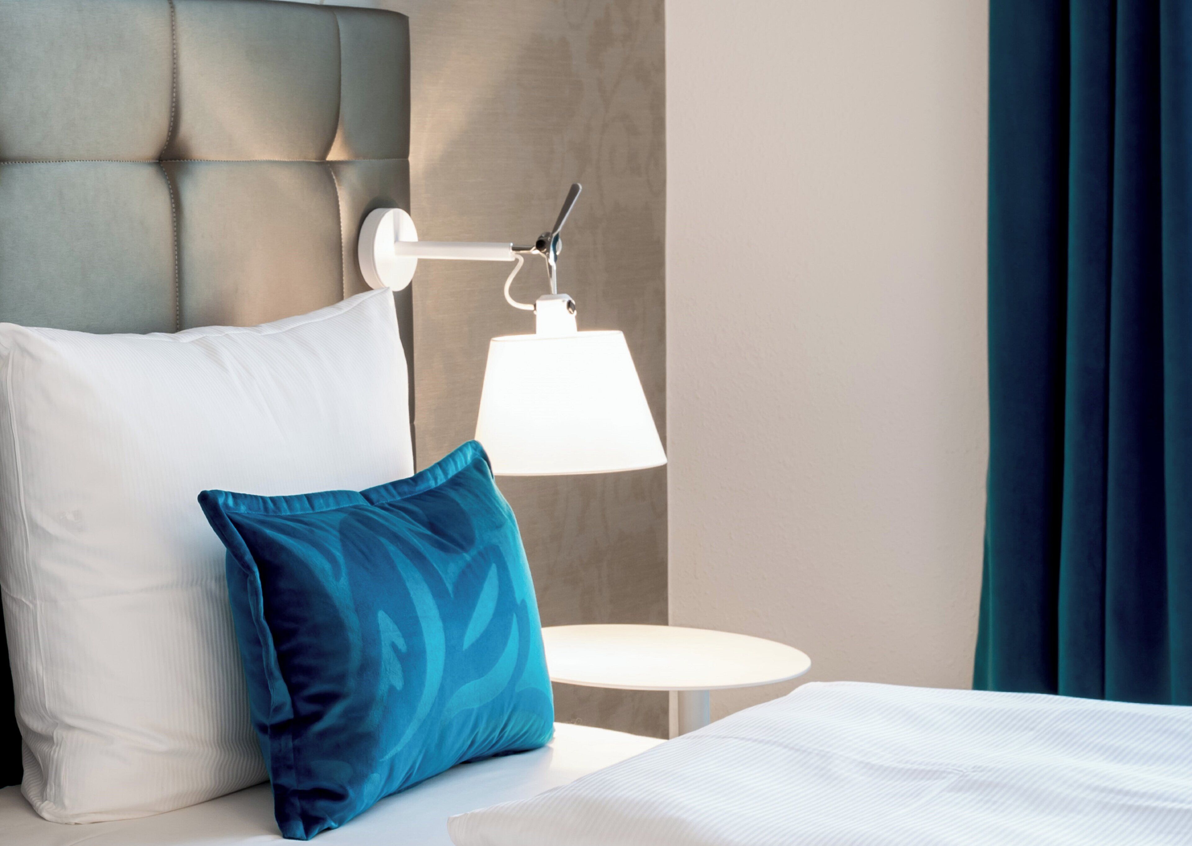 A hotel room with a bed, a pillow, a bedside table, a lamp, and a view of the outside.