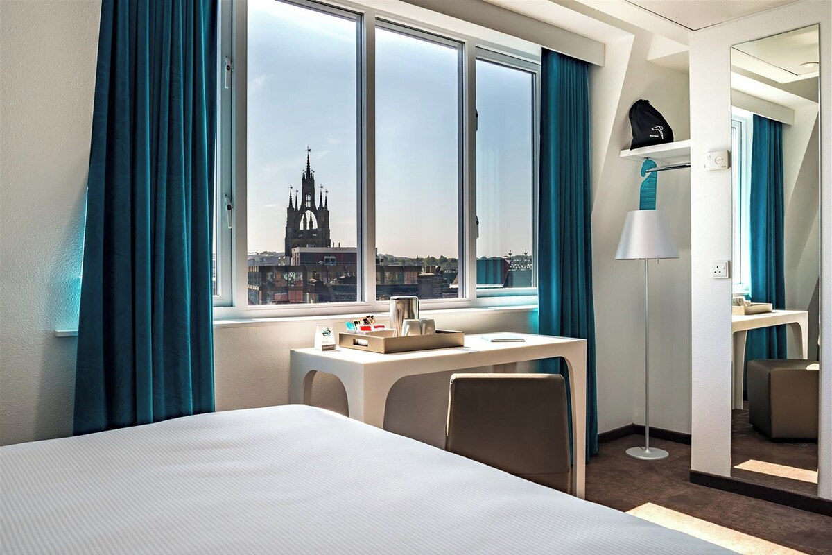 A hotel room with a bed, a desk, a chair, a lamp, and a view of a church through the window.
