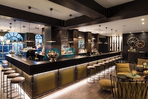 Zona de bar con una barra larga, taburetes y un área de estar con mesas redondas y sillas.