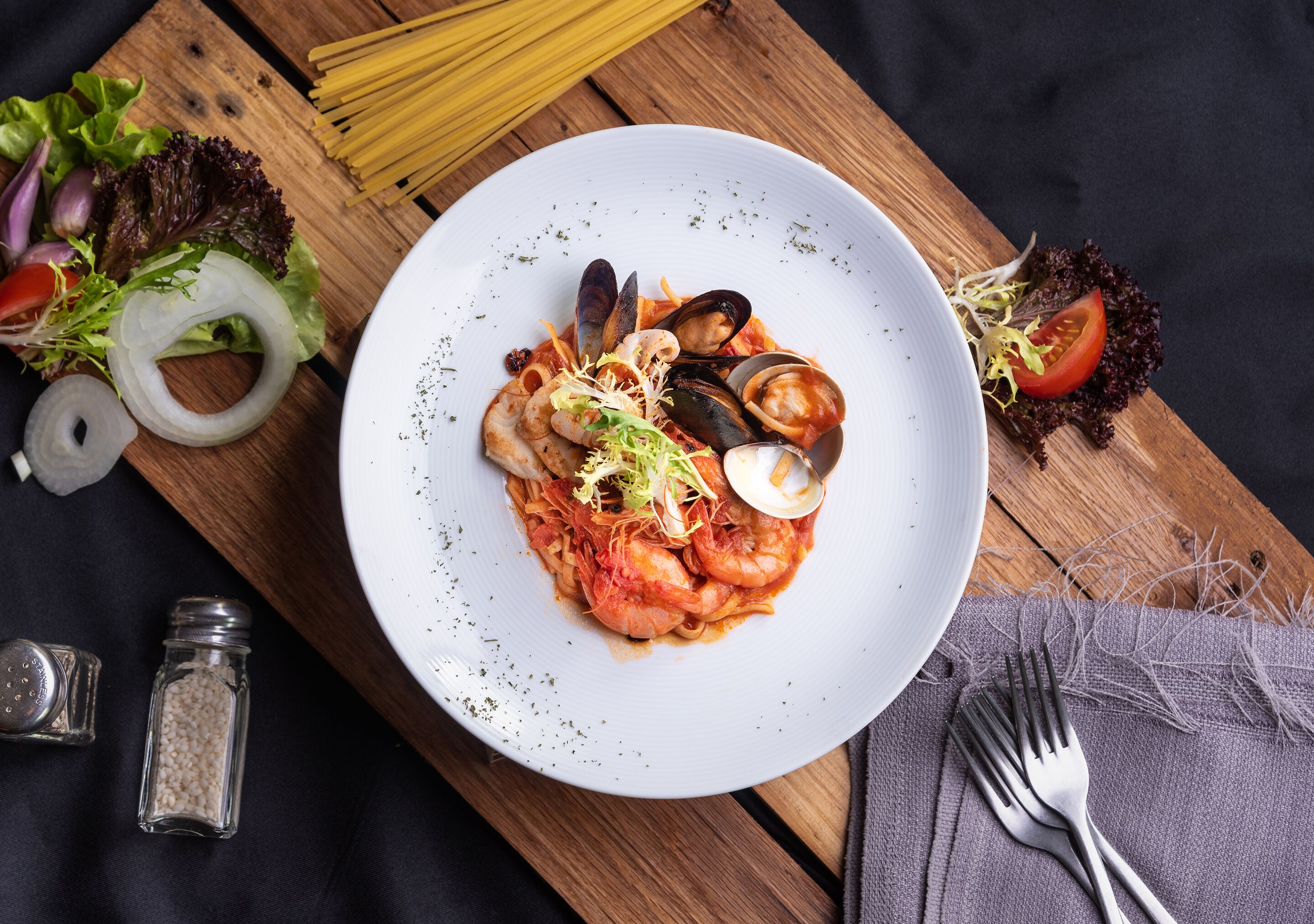 Un piatto di pasta di mare con cozze e gamberi, circondato da pasta, verdure e una forchetta su un tavolo di legno.