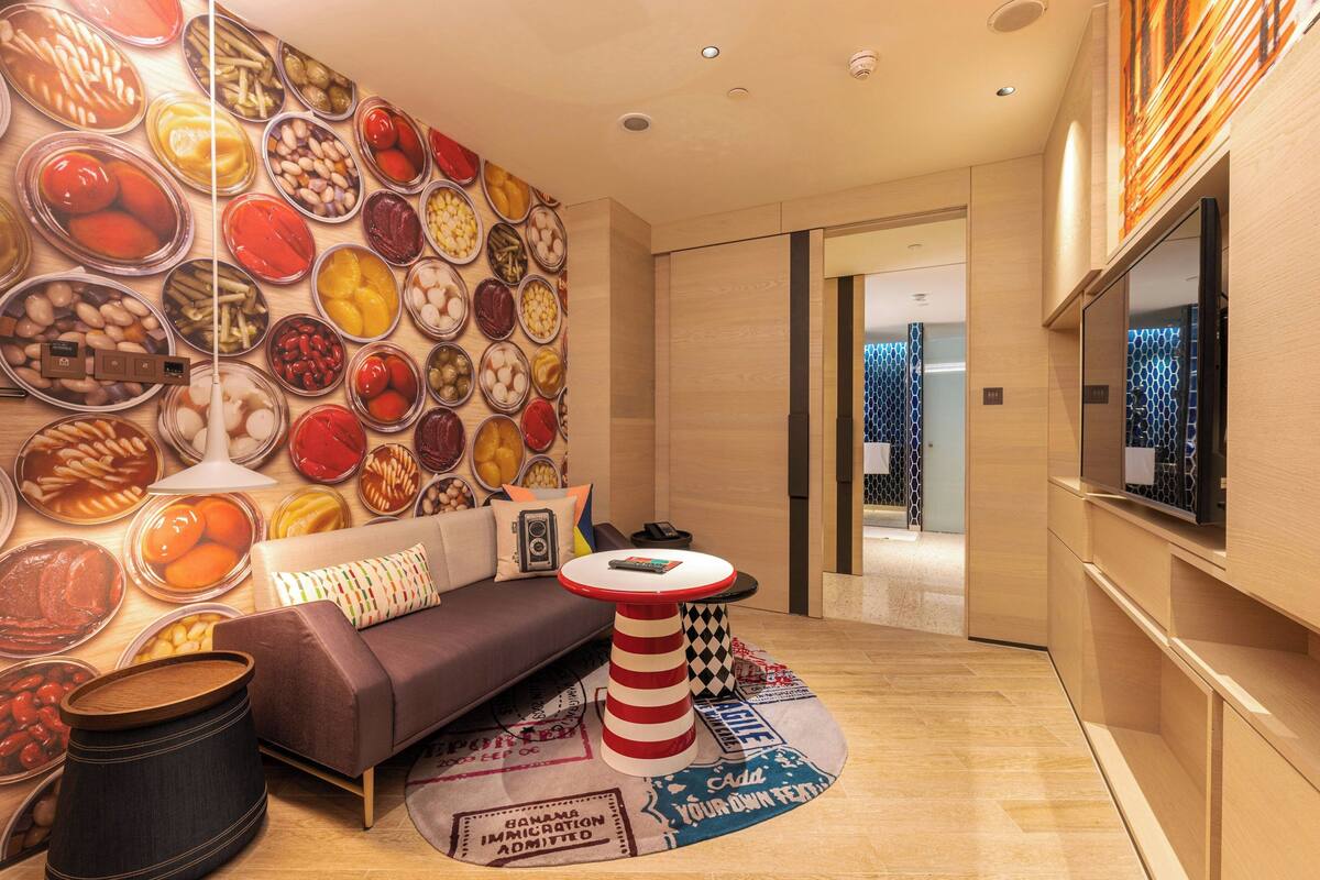 Une chambre d’hôtel moderne avec un grand mur décoré de corbeilles remplies de fruits et de légumes, un canapé, une table à rayures et un téléviseur.