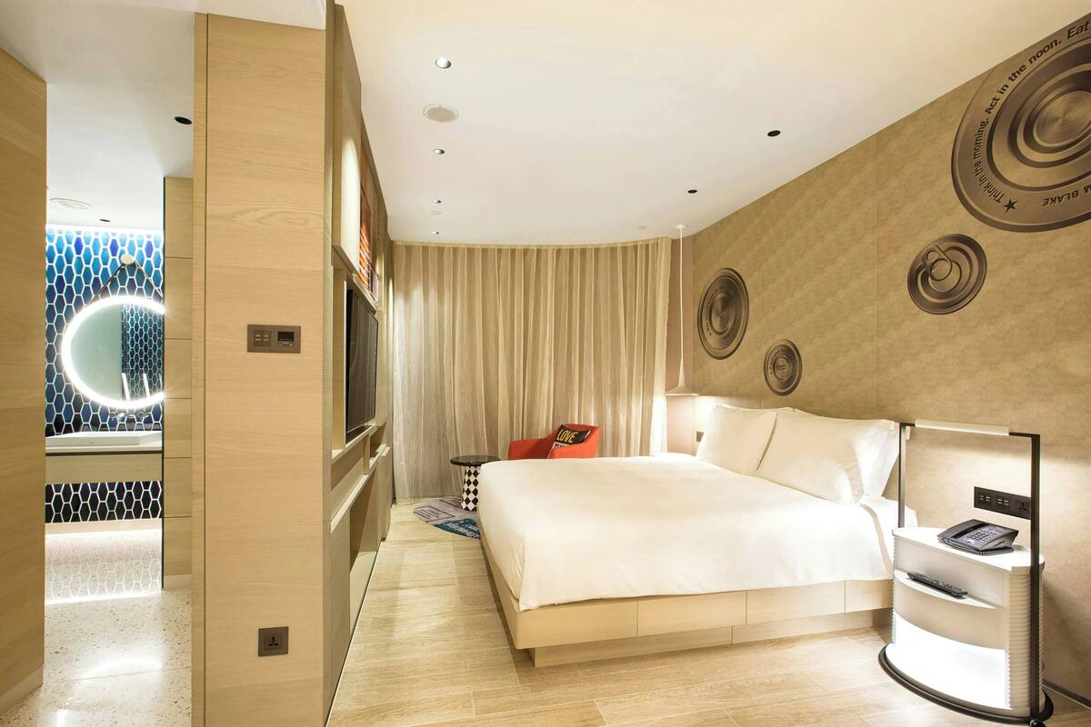 Une chambre d’hôtel moderne avec un grand lit, une table de chevet avec une lampe, un téléviseur fixé au mur et un rideau.