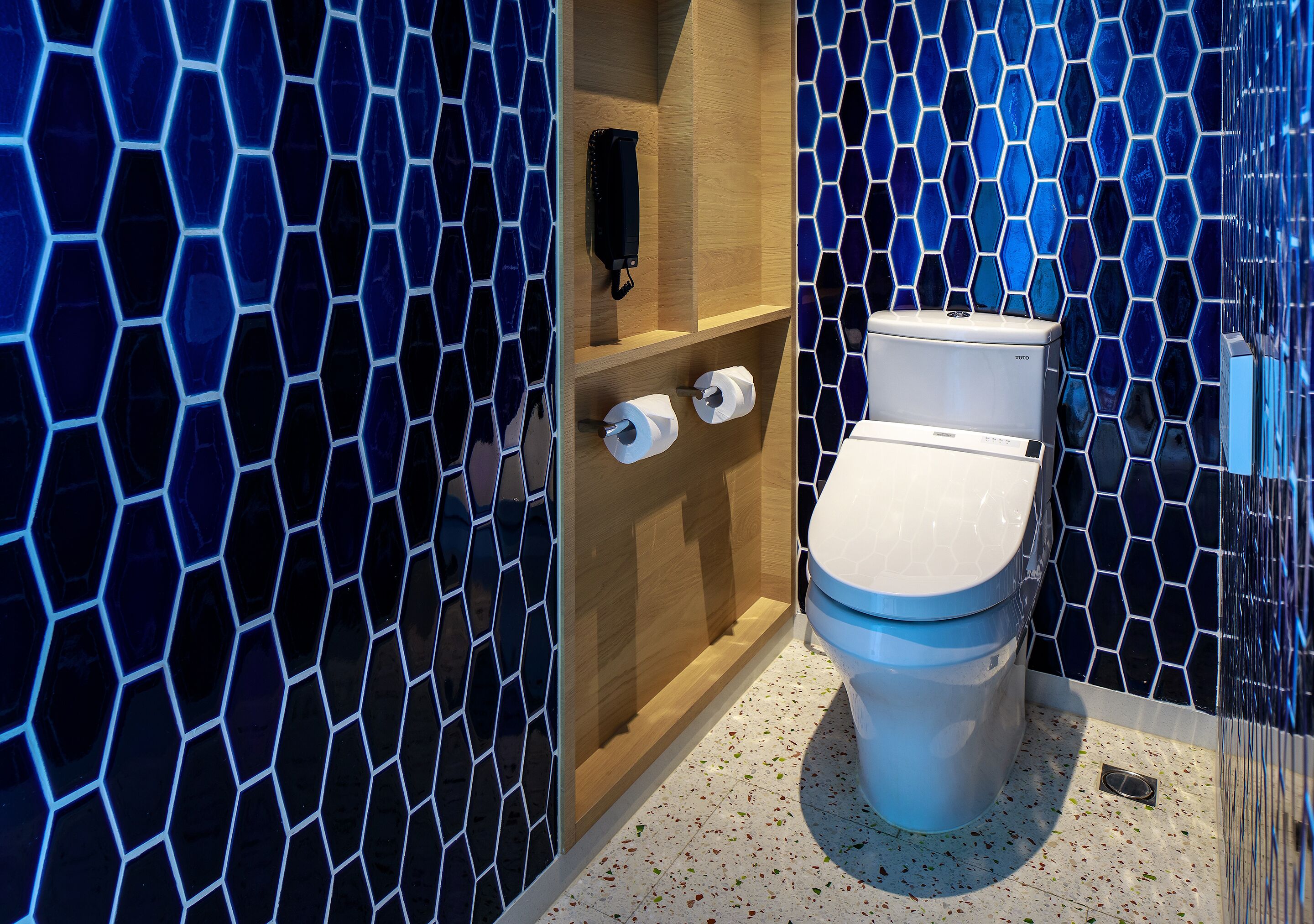 A modern bathroom with a hexagonal tile pattern, a toilet, and a wooden shelf with a black object and two rolls of toilet paper.