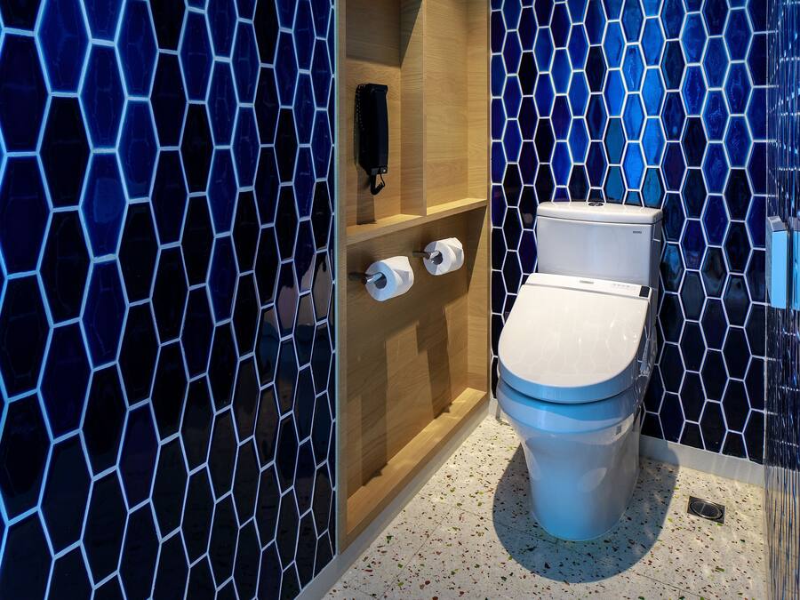 A modern bathroom with a hexagonal tile pattern, a toilet, and a wooden shelf with a black object and two rolls of toilet paper.
