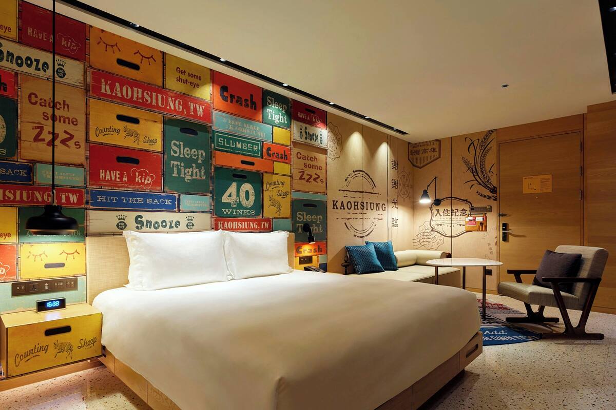 Une chambre d’hôtel moderne, équipée d’un lit, d’une petite table, d’une chaise et d’un mur décoré d’enseignes au style rétro.