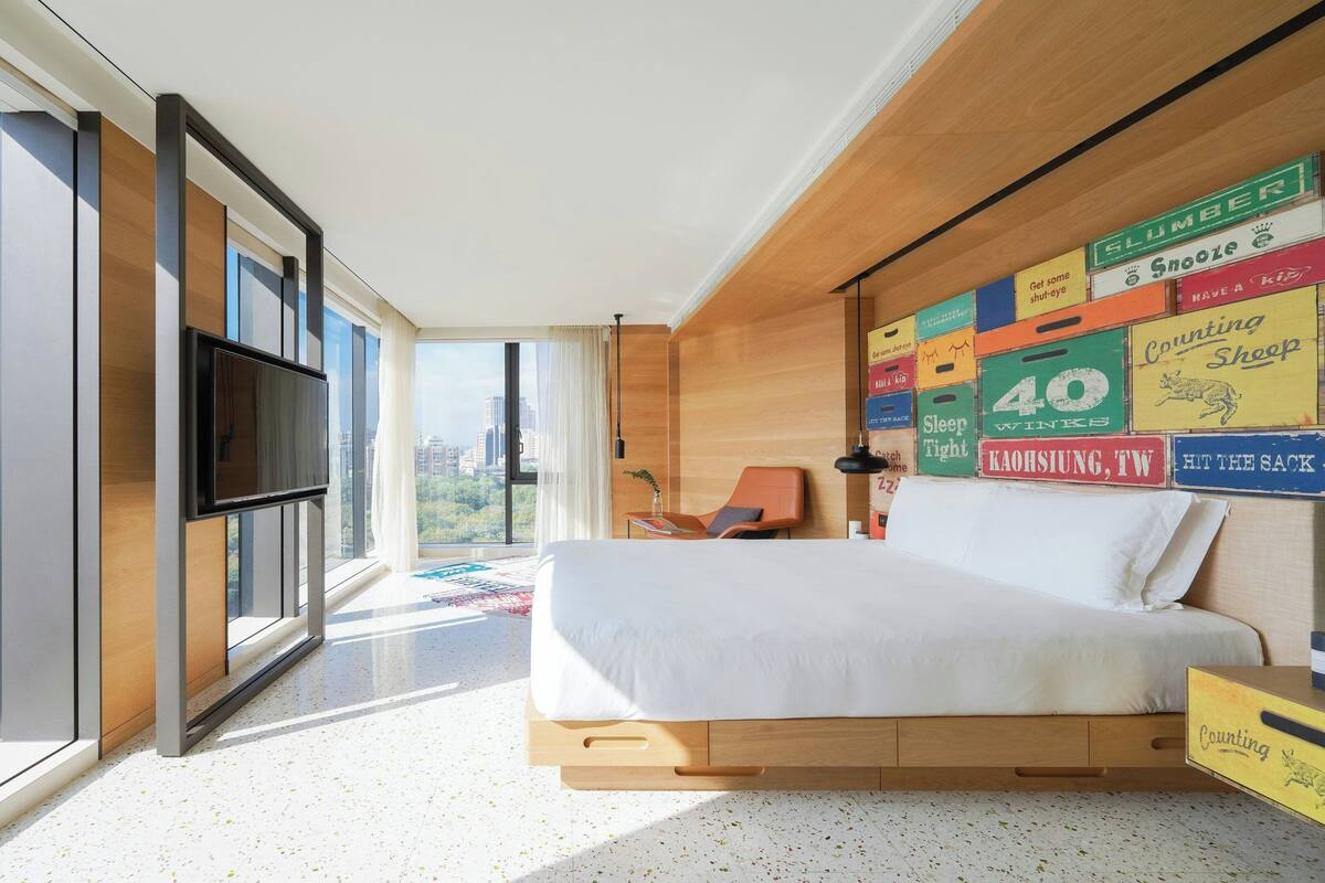 Une chambre moderne avec vue sur la ville, un téléviseur fixé au mur et un lit doté d’une tête de lit en bois.