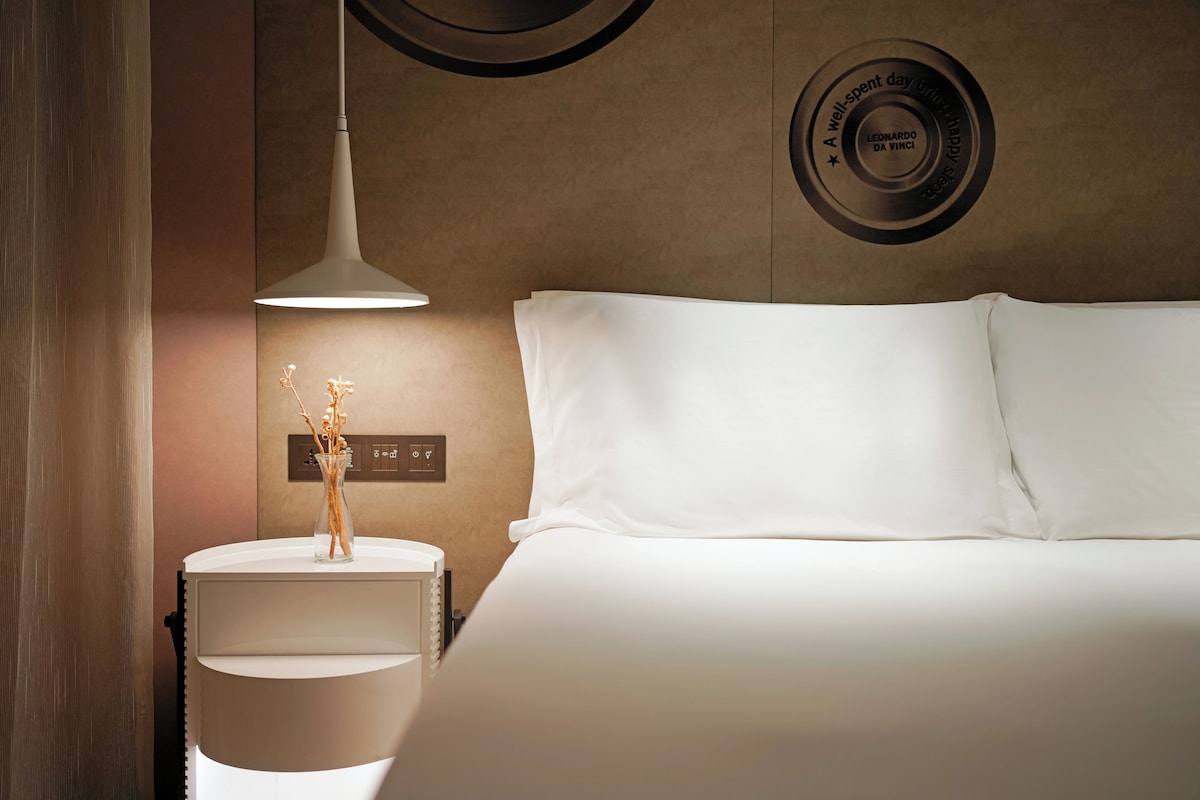 Hotellihuone, jossa on sänky, yöpöytä maljakolla, seinäkello ja moderni riippuvalaisin.