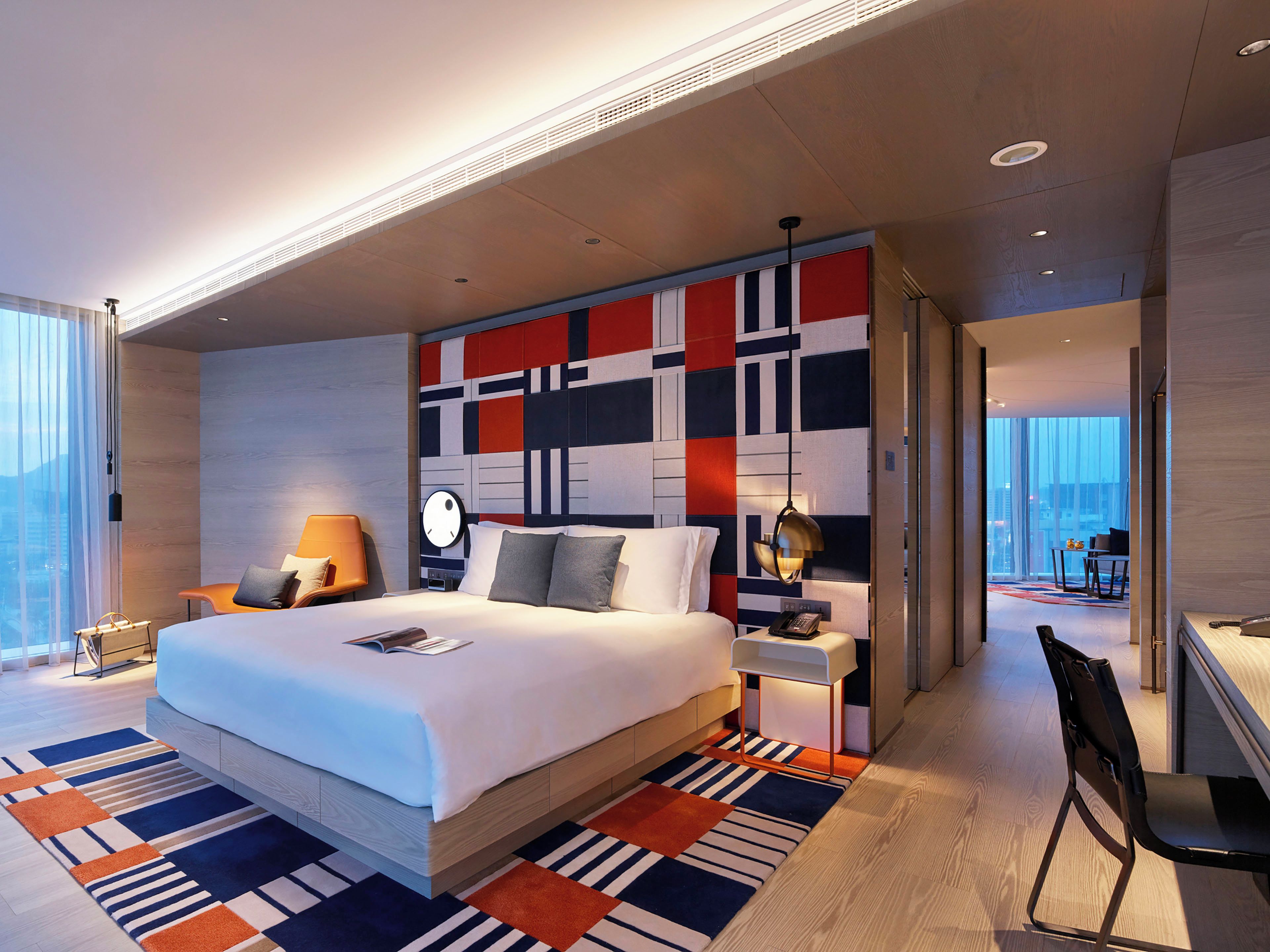 A modern hotel room with a large bed, a bedside table with a lamp, a chair, and a view of the city through large windows.