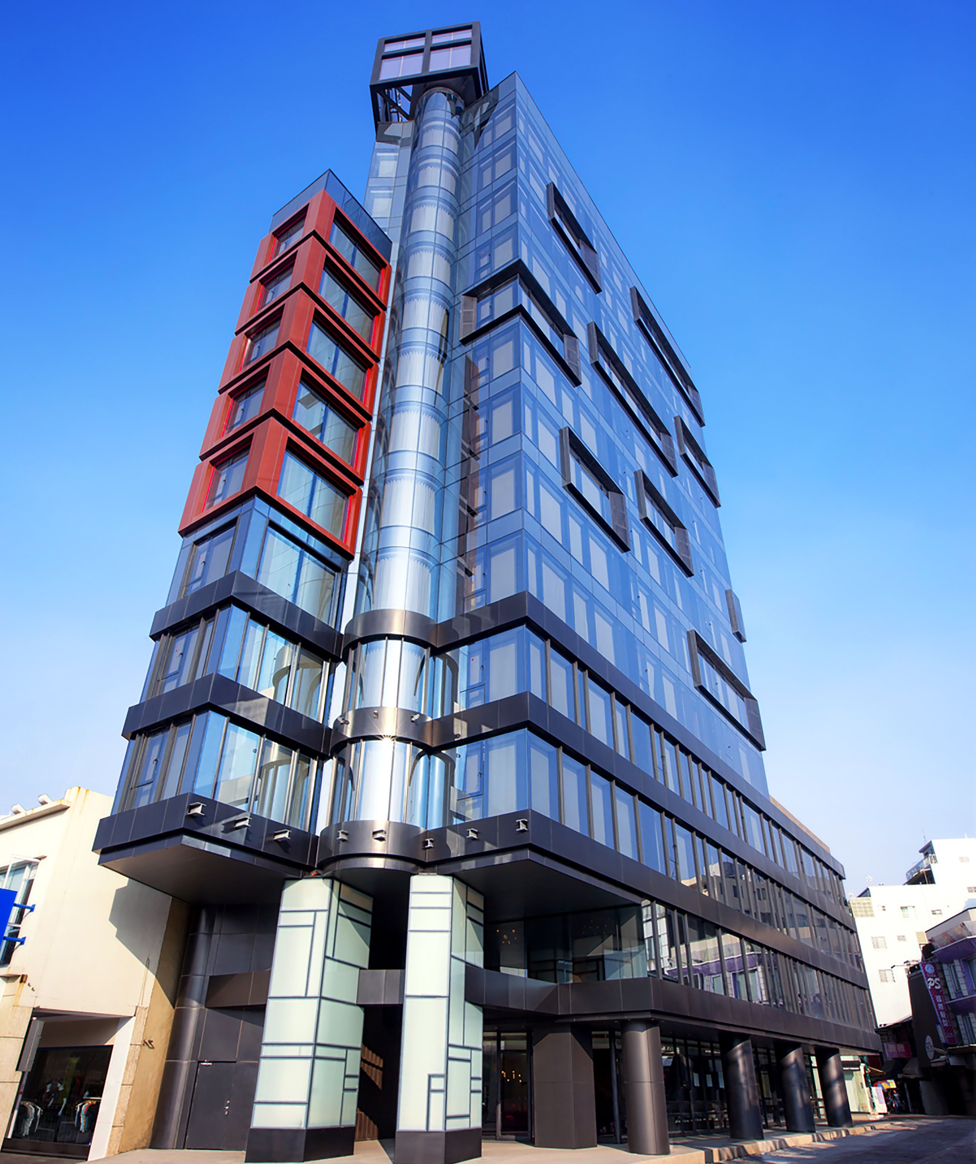 Un edificio moderno de varios pisos con fachada de vidrio, un marcado acento rojo en el lado izquierdo y un elemento de diseño curvo.