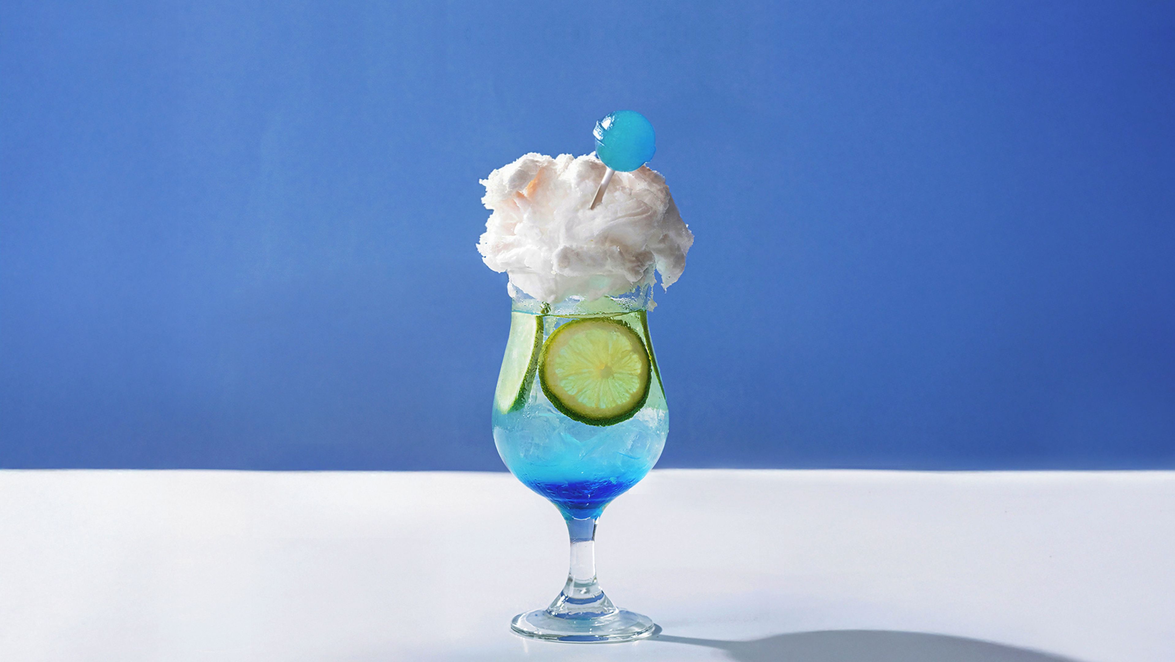 A cocktail glass with a blue liquid, a lime slice, and a blue spherical garnish on a white surface against a blue background.