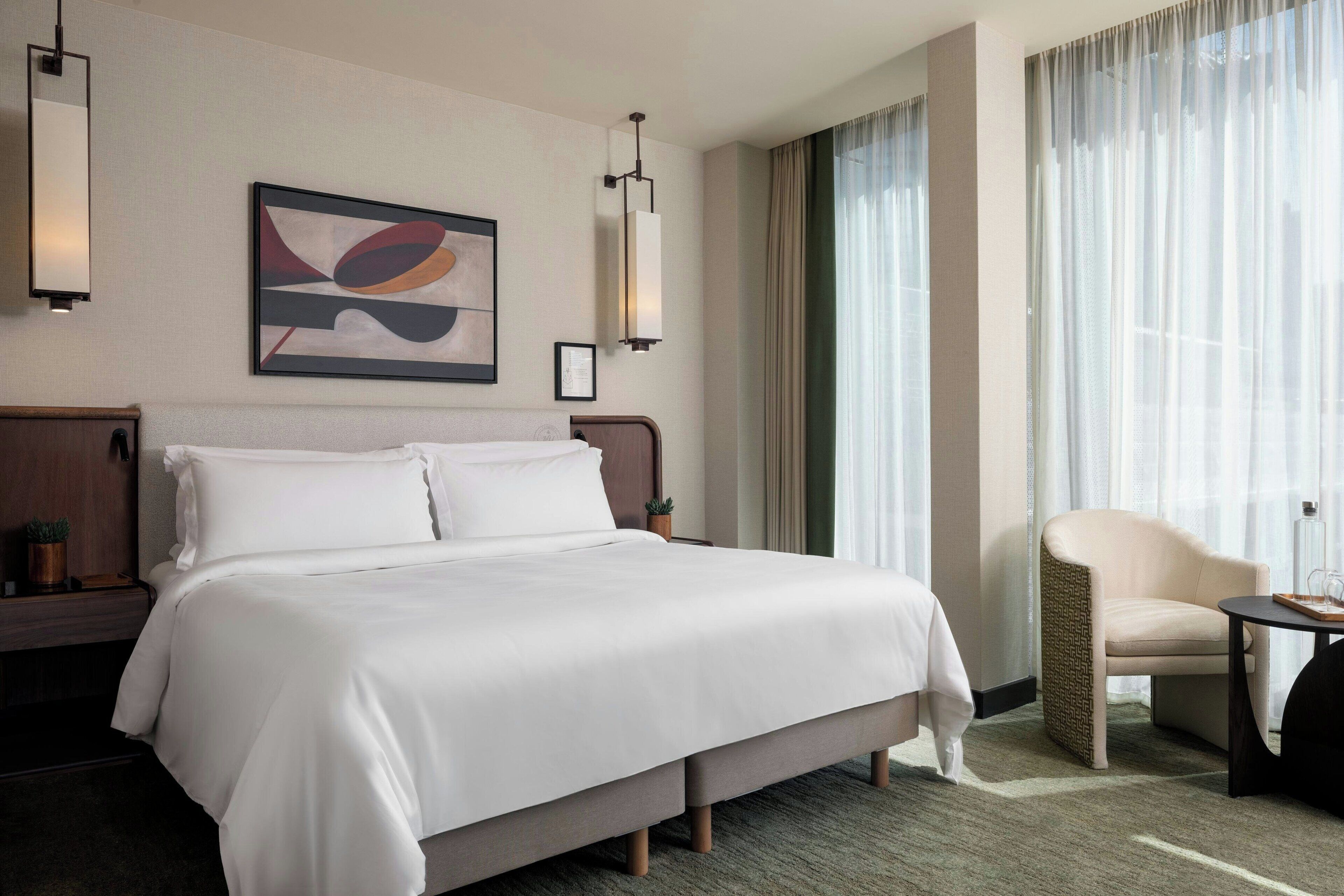 A hotel room with a large bed, a chair, a small table, and a painting on the wall.