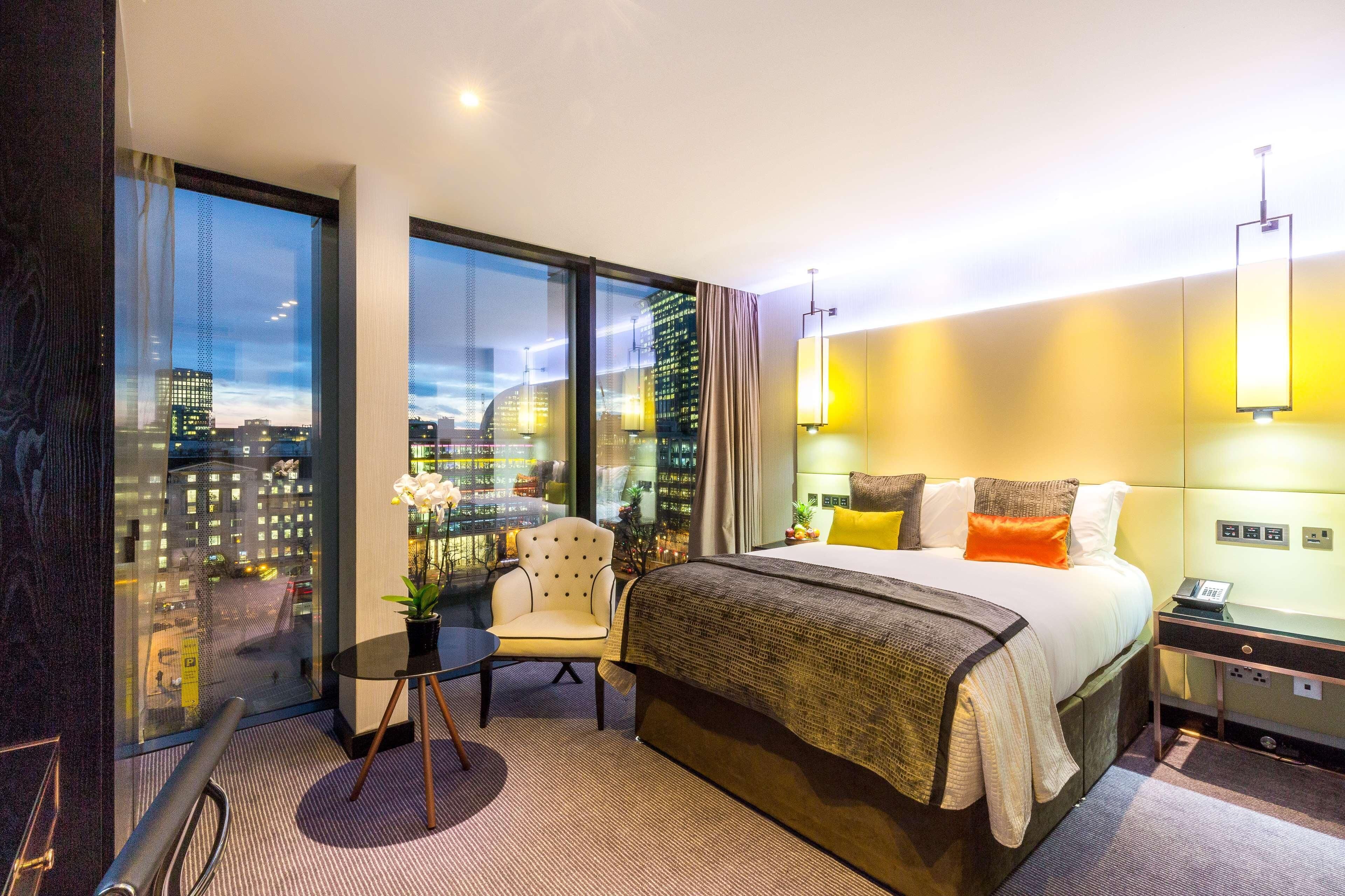 Habitación de hotel con una cama grande, una silla, una mesita y vistas de la ciudad a través de amplias ventanas.
