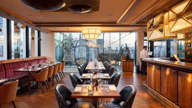 A restaurant with a long dining table, cushioned seating, and a bar area. - Montcalm Royal London House, Autograph Collection (London)