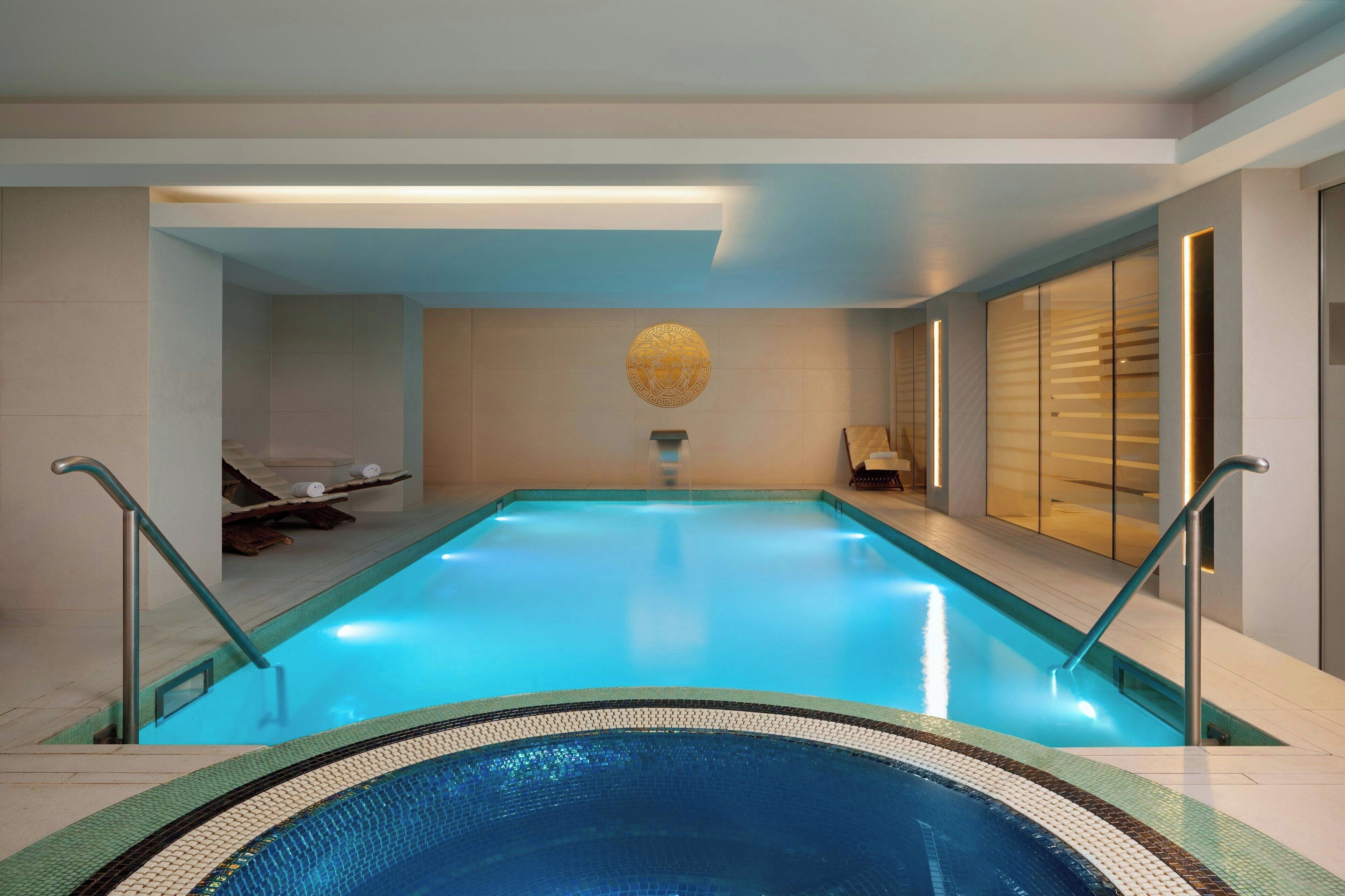 An indoor swimming pool with clear blue water, a central decorative wall, and a sunlit area with a bench and towel.