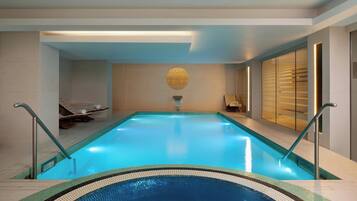 An indoor swimming pool with clear blue water, a central decorative wall, and a sunlit area with a bench and towel.
