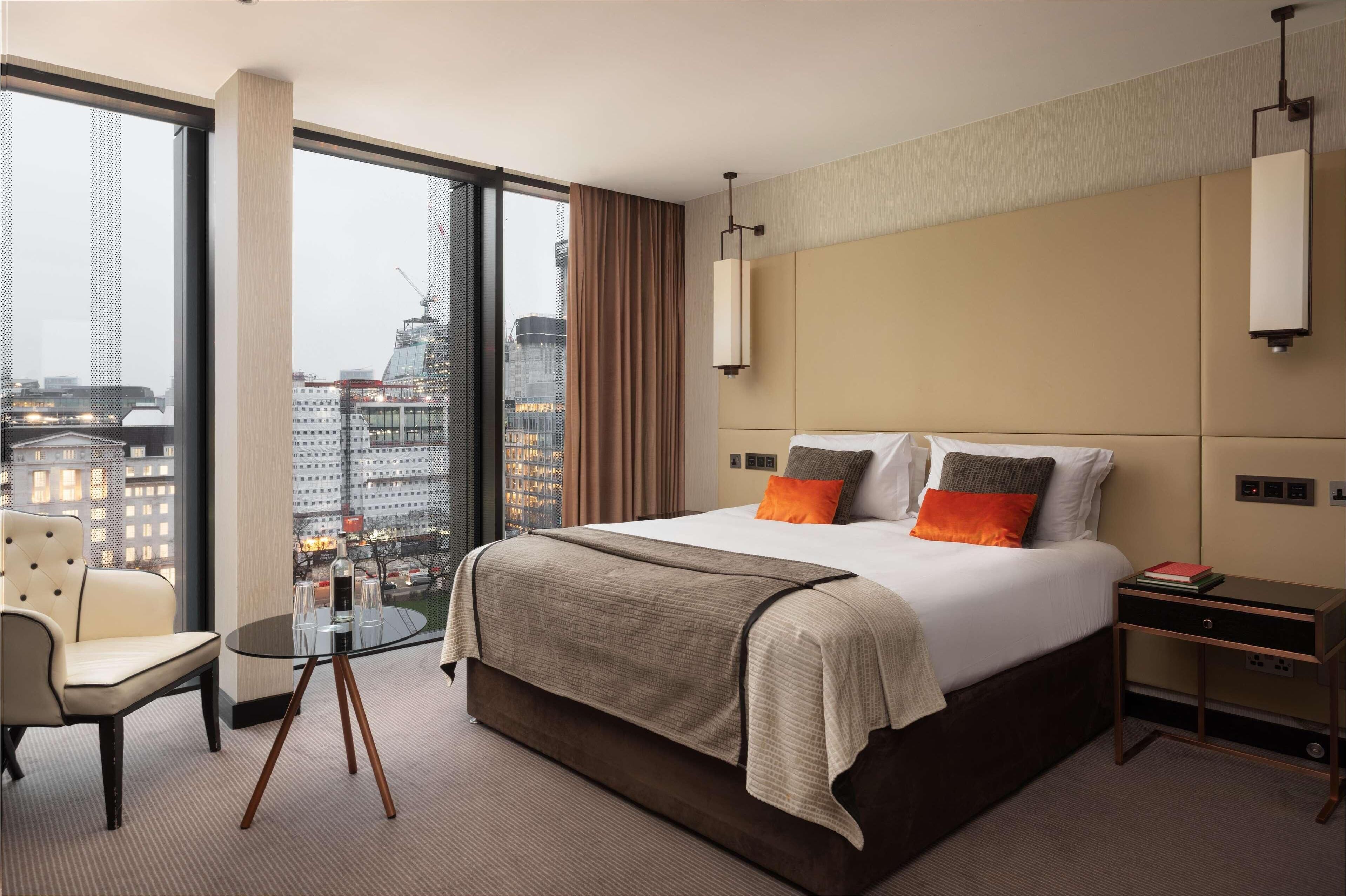 Habitación de hotel con una cama grande, una silla, una mesita y vistas a un paisaje urbano a través de amplias ventanas.