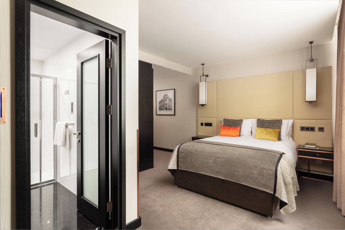 Habitación de hotel con cama, mesitas de noche, una imagen enmarcada en la pared y una puerta que da al baño.