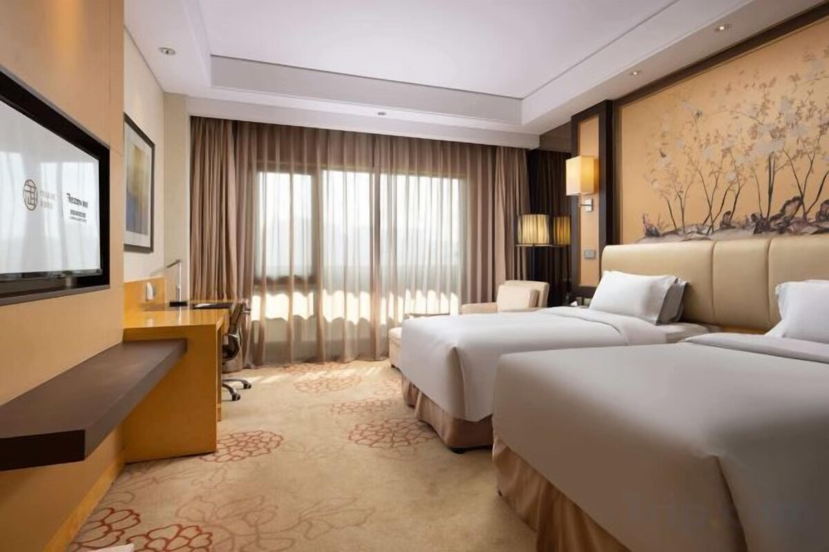 A hotel room with two beds, a desk, a television, and a large window with curtains.