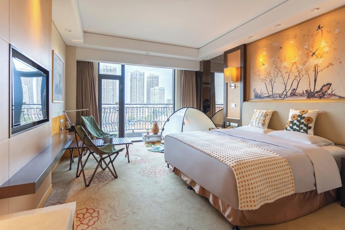 Une chambre d’hôtel avec un lit, un bureau, une chaise, un balcon donnant sur des immeubles de grande hauteur et un tableau accroché au mur.