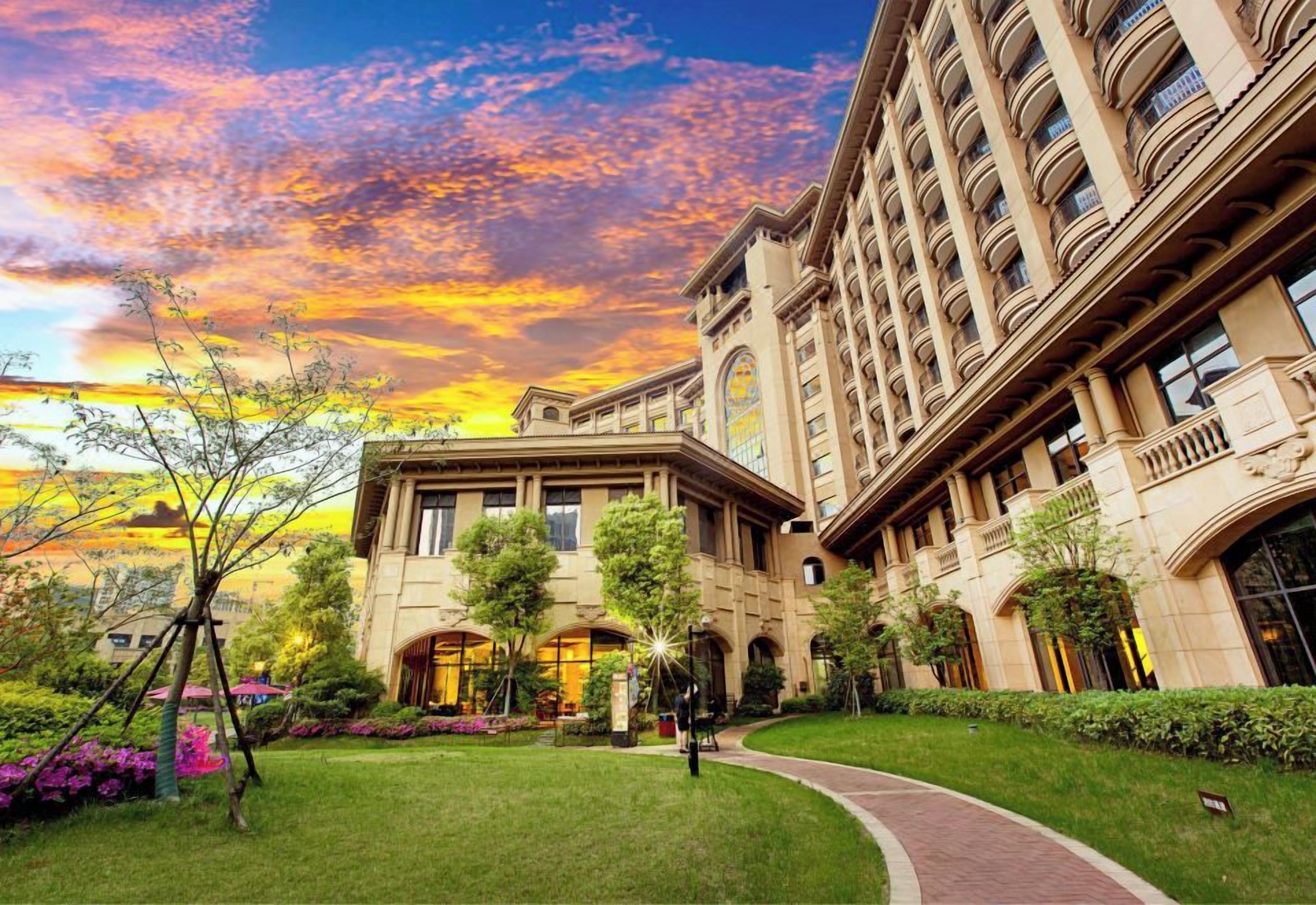 A grand hotel with a classical architectural style, featuring a central courtyard with a pathway, surrounded by lush greenery and a large building with multiple balconies.