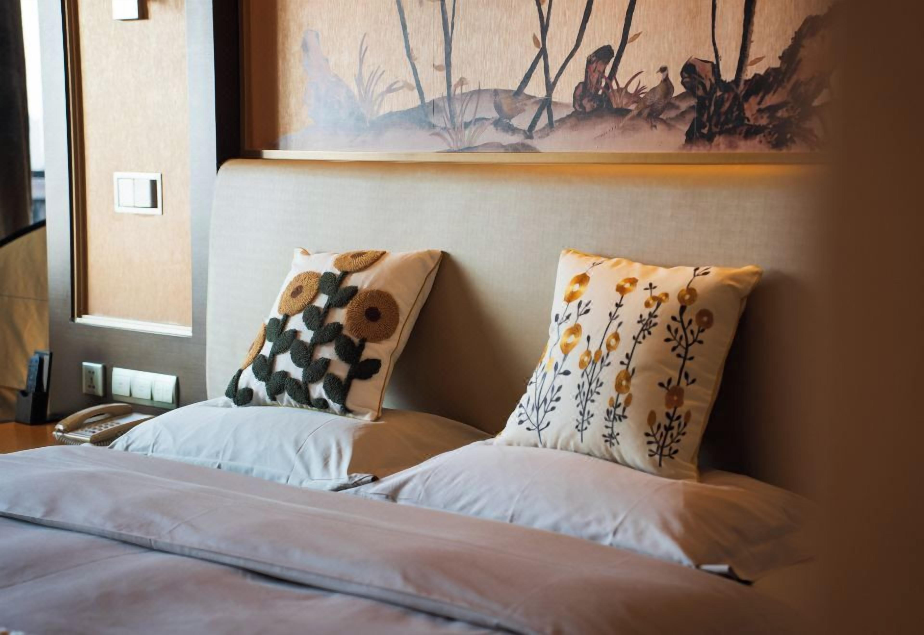 Habitación de hotel con una cama, dos almohadas con motivos florales, un cuadro en la pared y un teléfono en la mesita de noche.