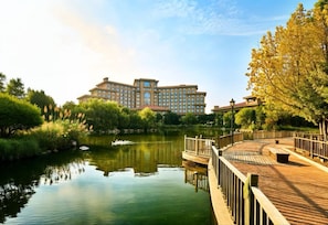 A large hotel with a reflective water body in front, surrounded by greenery and a wooden deck.