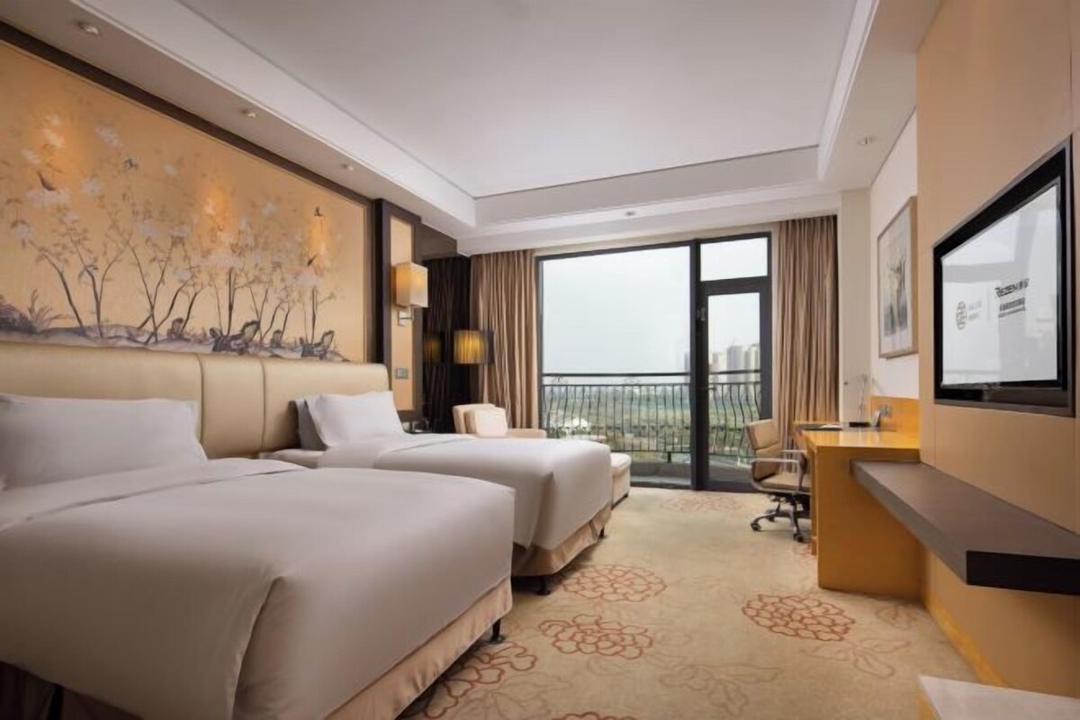 Une chambre d’hôtel avec deux lits, un bureau, une chaise, une grande fenêtre et un tableau au mur.
