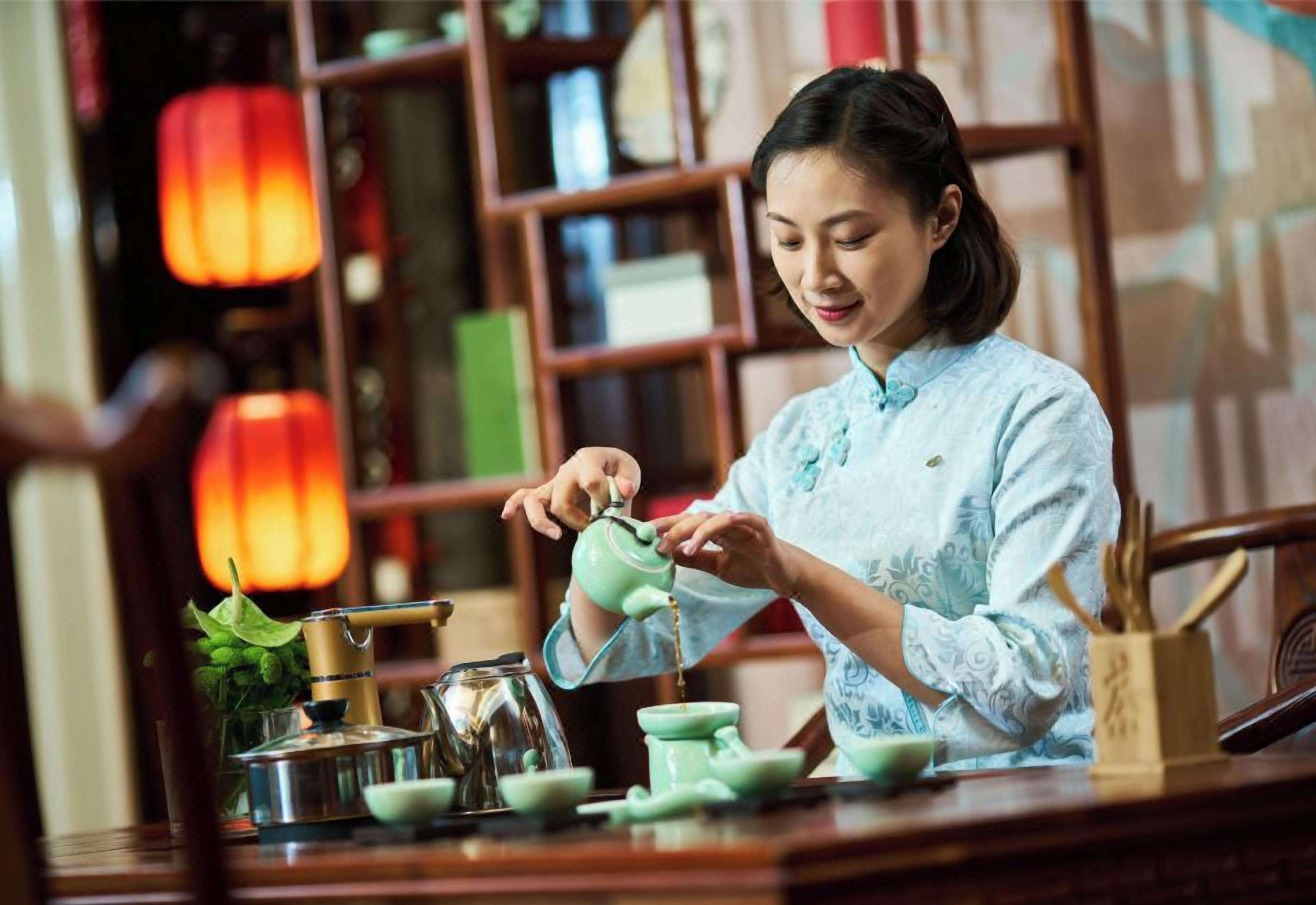 A person pouring tea from a teapot into a cup, with a tea set on a table, surrounded by books and a bookshelf in the background.