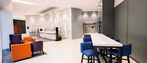 A modern hotel lobby with a long dining table, blue chairs, orange sofas, and a reception area in the background.