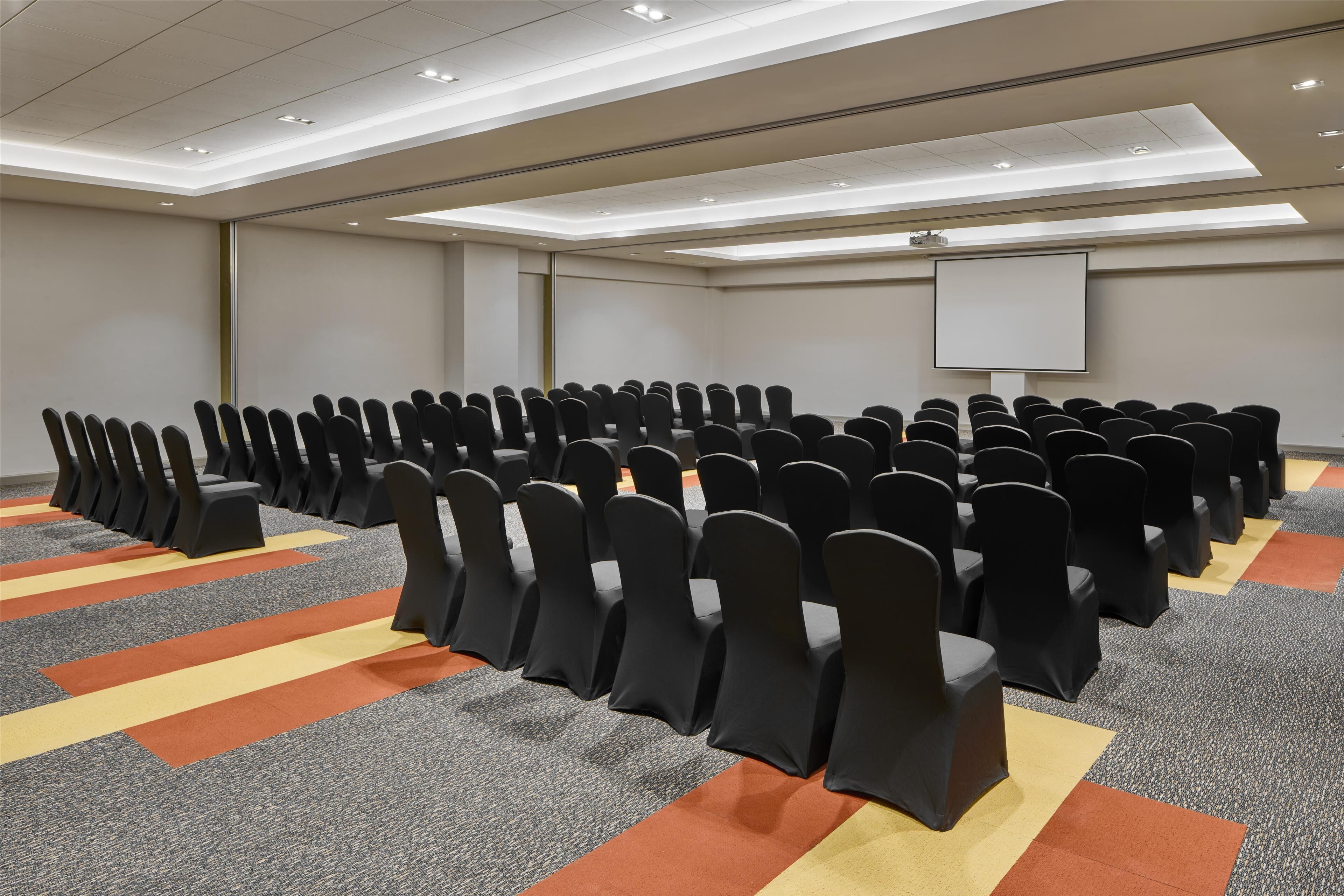 A conference room with rows of chairs, a projector screen, and a carpet with geometric patterns.