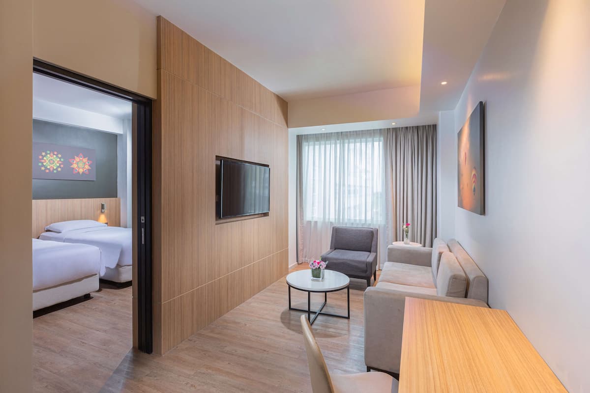 A modern hotel room with a wooden floor, a flat-screen TV mounted on the wall, a sofa, a small round coffee table with a vase of flowers, and a bed with white bedding.
