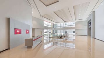 Eine großzügige Lobby mit modernem Design, inklusive einer Rezeption, Glastrennwand und einer zentralen Treppe.