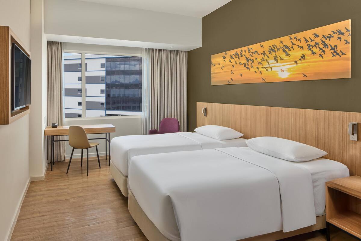 A hotel room with two beds, a desk, a chair, and a large window with a view of buildings.