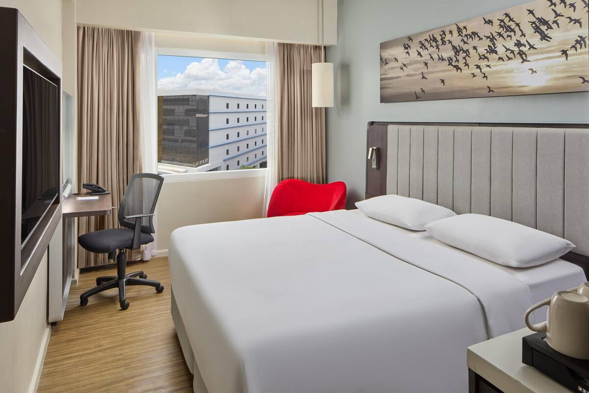 A hotel room with a bed, a desk with a chair, a TV, and a window with curtains.