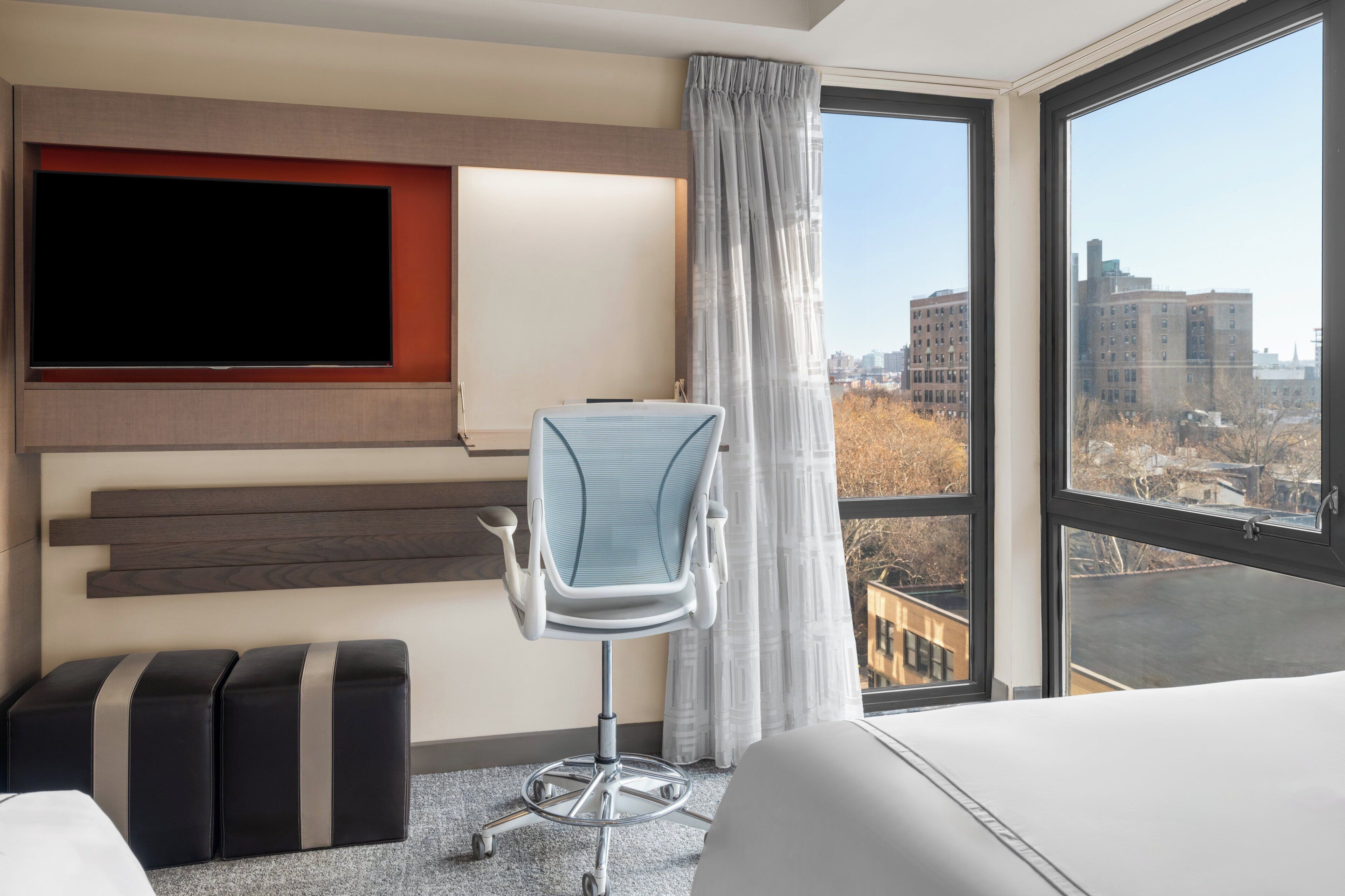 A hotel room with a large window, a flat-screen TV mounted on the wall, a modern office chair, and a bed.